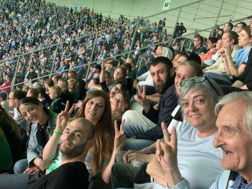 Menschen sitzen in den rängen eines ausverkauften Fußballstadions und lächeln freudig in die Kamera