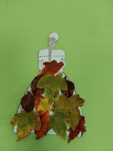 Eine Bastelarbeit, welche die Zeichnung einer Frau in einem Kleid zeigt. Das Kleid ist mit Herbstlaub beklebt.