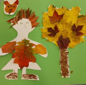 Eine Bastelarbeit, die ein Mädchen neben einem Baum zeigt, mit einem Haustier im Hintergrund. Das Haustier und der Baum sind aus Herbstlaub gestaltet. Das Kleid des Mädchens ist mit Herbstlaub beklebt.