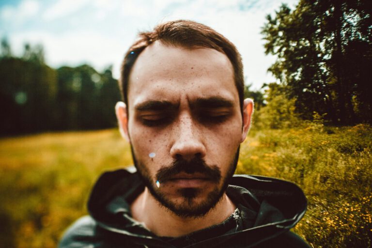 Trauriger Mann in Nahaufnahme. Er steht Mitten in einer sommerlichen Blumenwiese, und schließt traurig die Augen. Der Hintergrund ist verschwommen und unscharf.