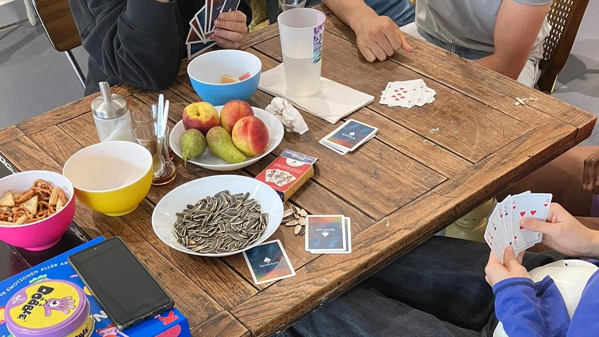 Kartenspiele und bunte Schüsseln mit Snacks stehen auf einem Holztisch. 3 Menschen sitzen an diesem Tisch und spielen ein Kartenspiel.