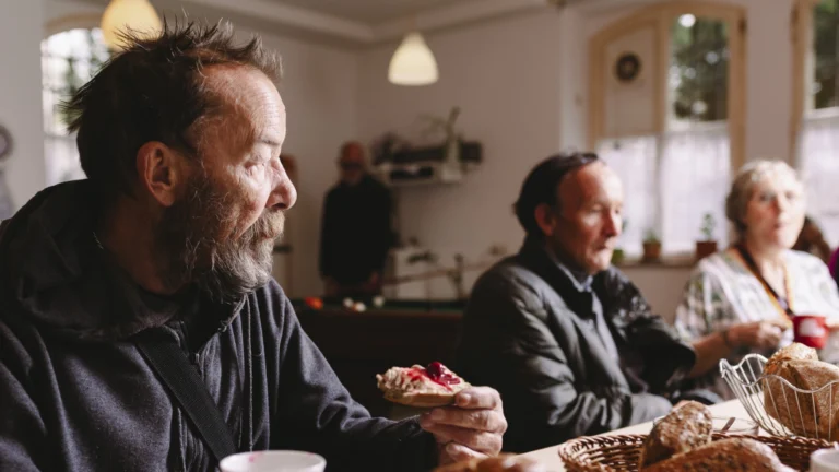 Drei Wohnungslose Menschen genießen ein Frühstück im Café Emmaus. Der Mann im Vordergrund hält ein Marmeladenbrot mit roter Marmelade in der Hand.