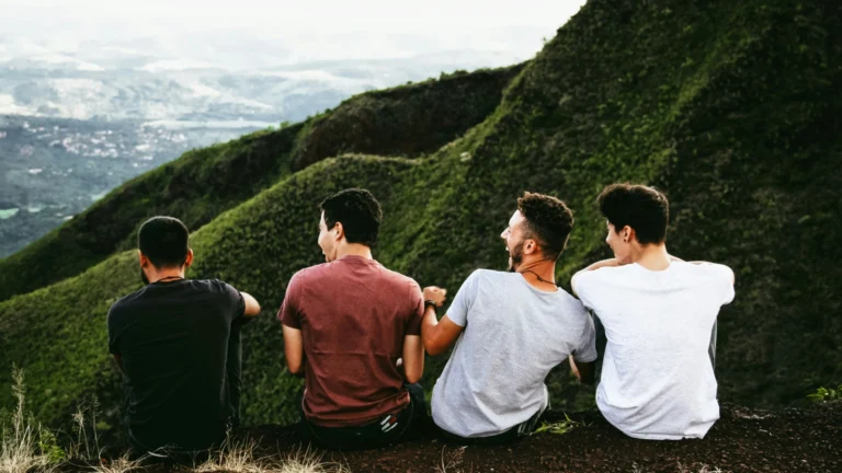 Eine Gruppe junger Männer sitzt auf einem Bergkamm und schaut herab ins Tal. Offenbar amüsieren oder freuen sie sich über etwas, das sie beobachten, das aber im Bild nicht zu sehen ist.
