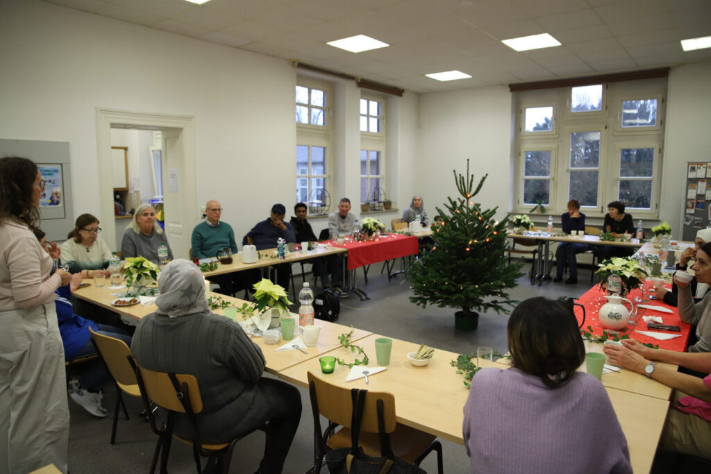 Viele Menschen sitzen an einer zu einem großen Quadrat aufgebauten Tafel. Vor ihnen auf den Tischen liegt weihnachtliche Dekoration. In der Mitte steht ein mit einer Lichterkette geschmückter Weihnachtsbaum