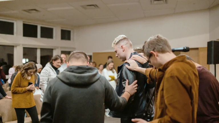 Eine Gruppe von Menschen gibt sich gegenseitig Kraft und Zuspruch. Ein junger Mann mit blonden Haaren steht mit gesenktem Blick in der Mitte. Neben und hinter ihm stehen weitere Männer, die ihm die Hand auf die Schulter legen