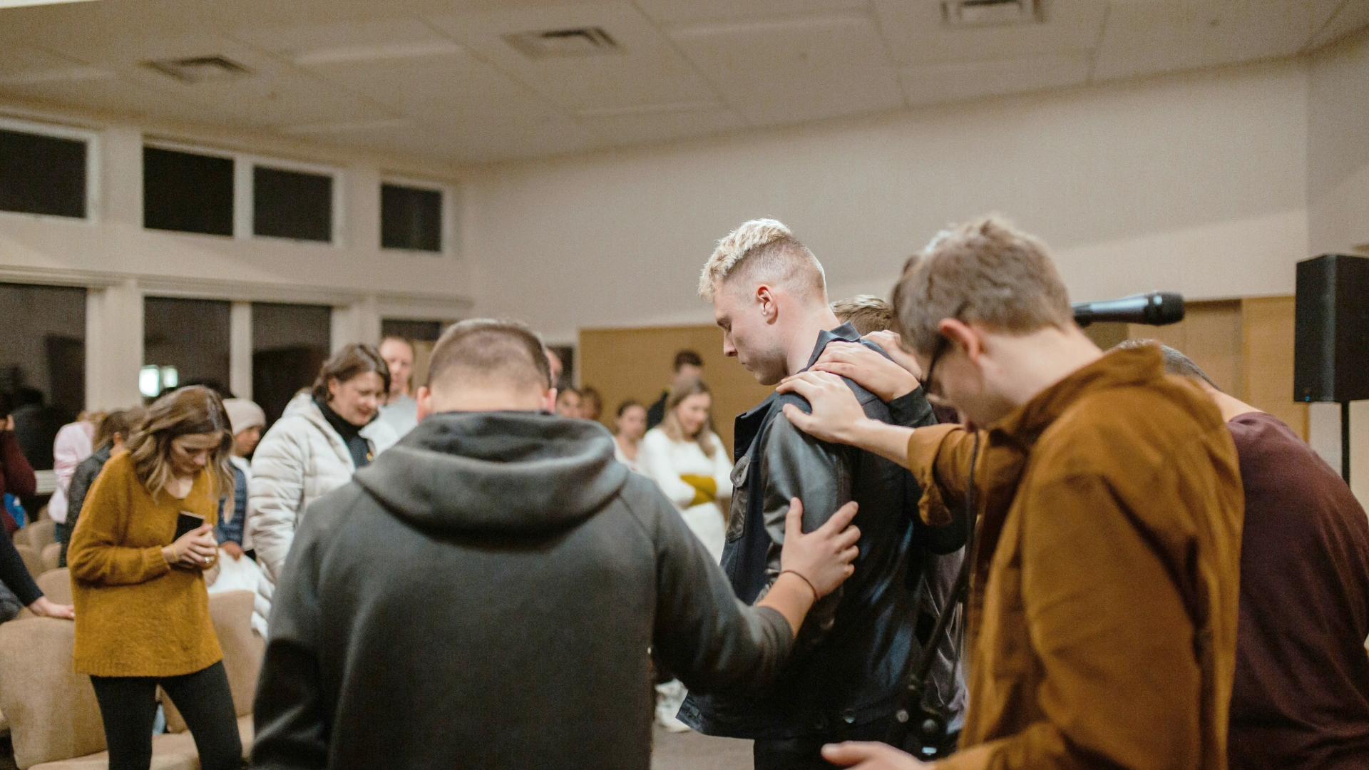Eine Gruppe von Menschen gibt sich gegenseitig Kraft und Zuspruch. Ein junger Mann mit blonden Haaren steht mit gesenktem Blick in der Mitte. Neben und hinter ihm stehen weitere Männer, die ihm die Hand auf die Schulter legen